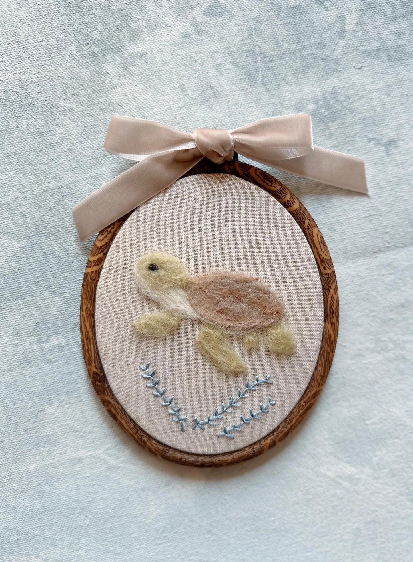 Felted turtle on fabric with wooden frame and pink ribbon on textured light blue background
