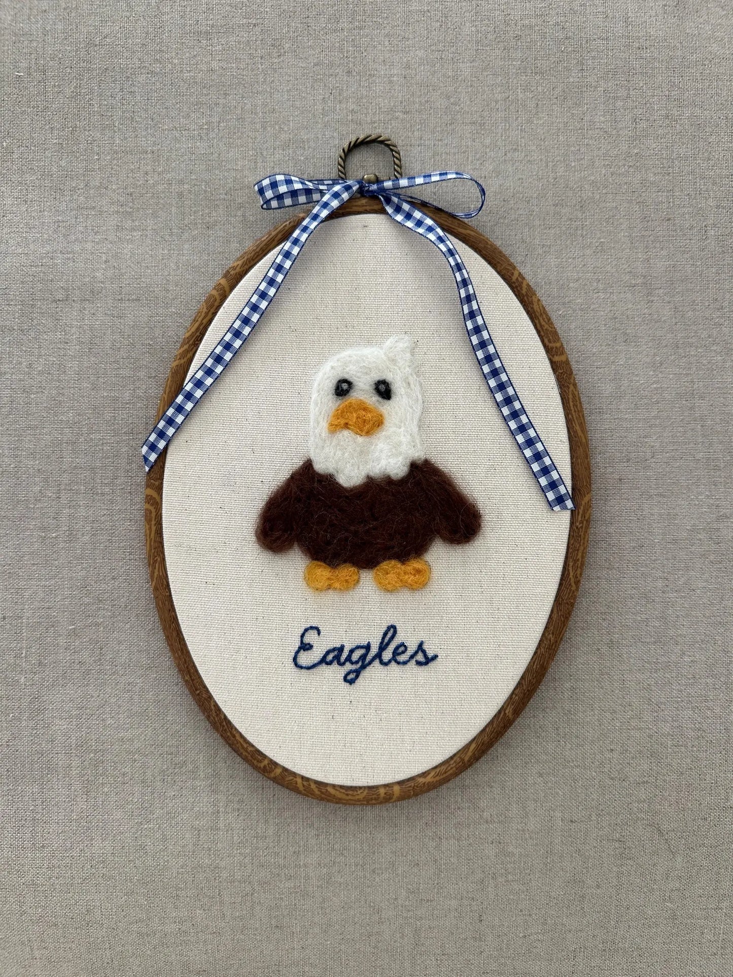 Embroidered eagle design on a wooden hoop with a blue and white checkered ribbon against a beige background.