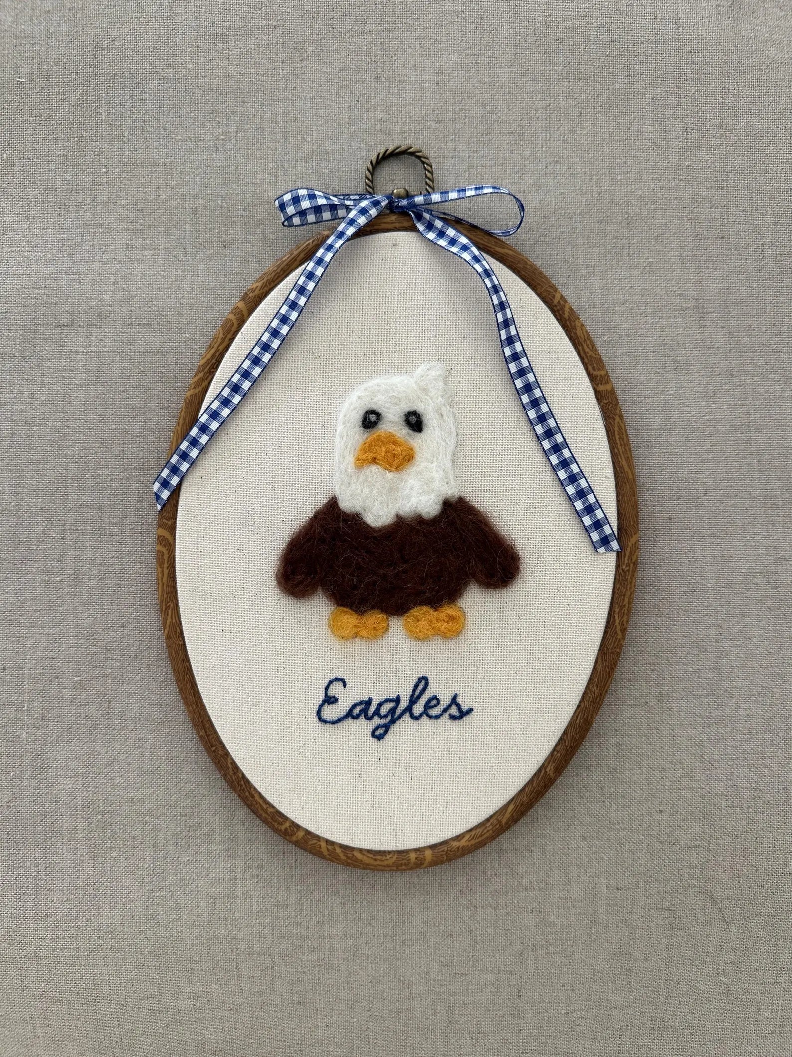 Embroidered eagle design on a wooden hoop with a blue and white checkered ribbon against a beige background.