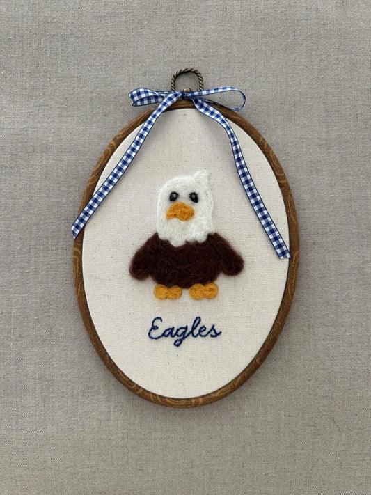 Embroidered eagle design on a wooden hoop with a blue and white checkered ribbon against a beige background.