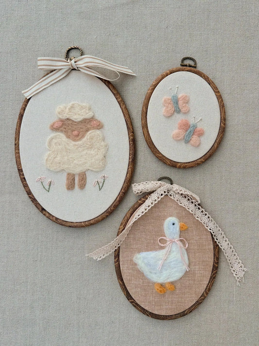 Three embroidered hoop art pieces featuring a sheep, duck, and butterflies on a textured fabric background.