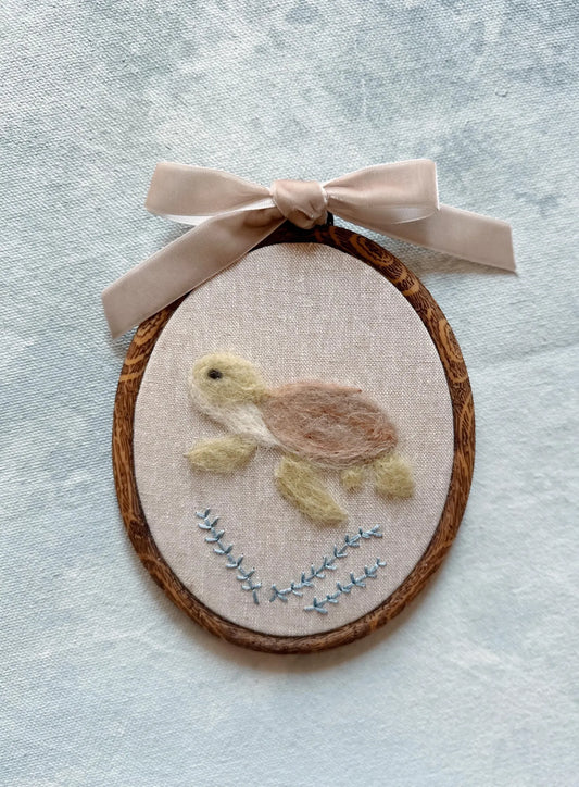 Felted turtle on fabric with wooden frame and pink ribbon on textured light blue background