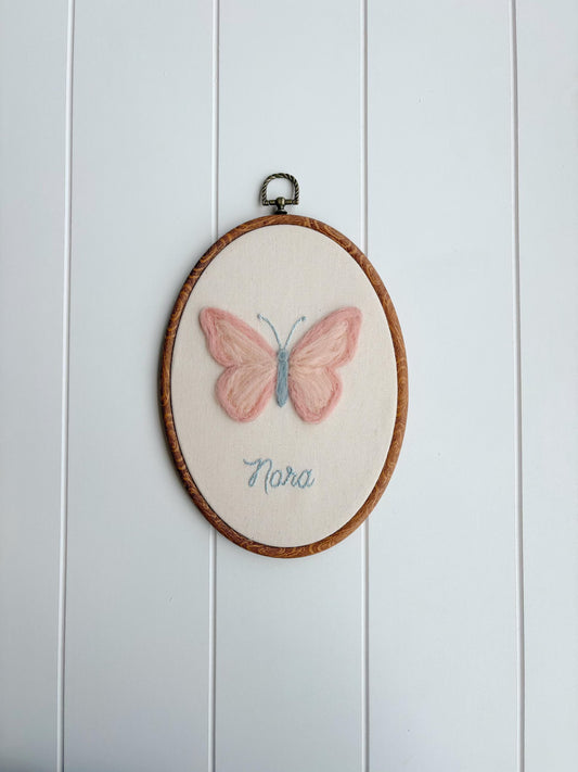 Embroidery hoop with a pink butterfly design and 'Nora' text on a white wooden panel background
