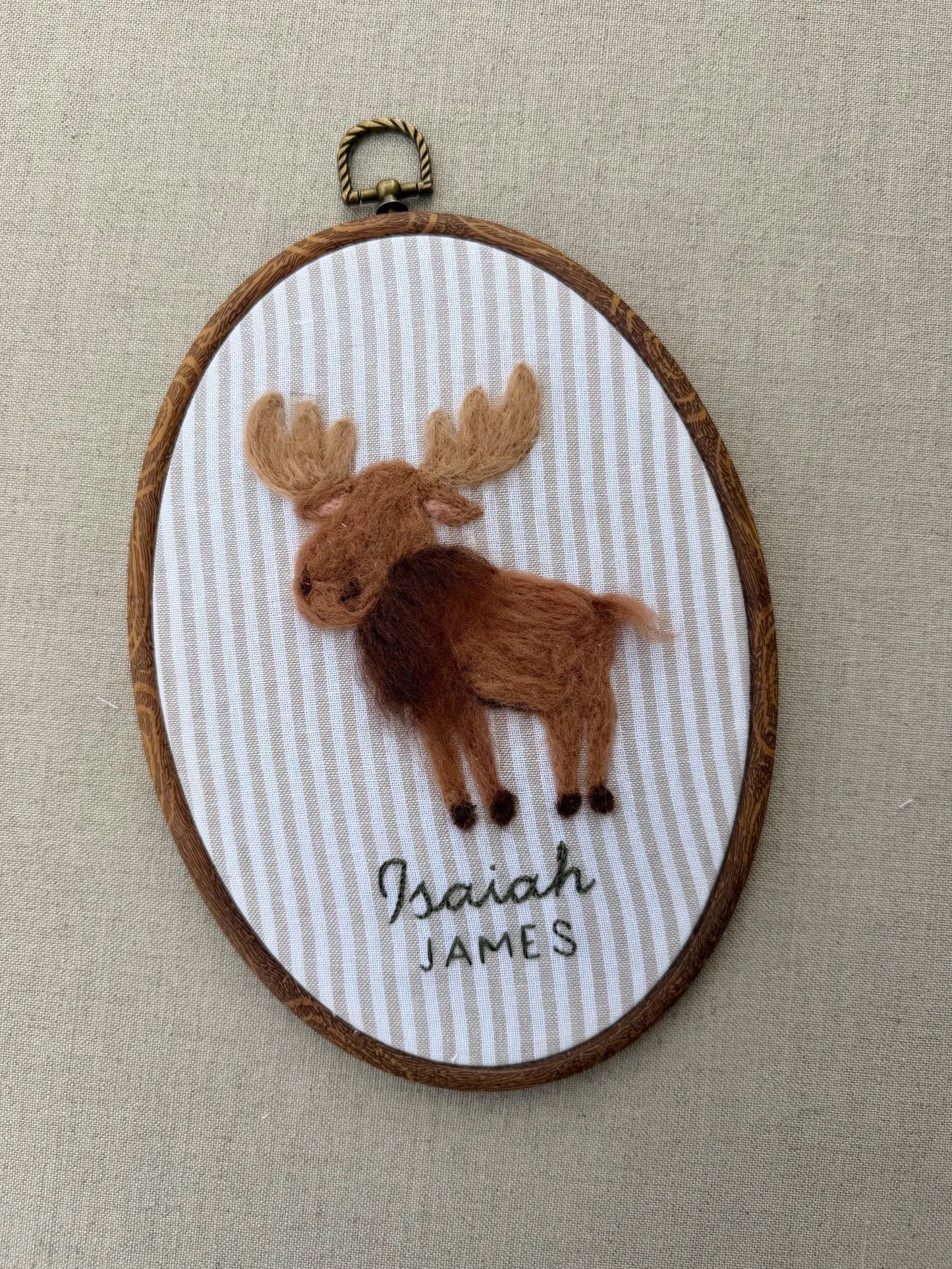Felted moose design with 'Isaiah James' text in a wooden hoop on a striped fabric background.