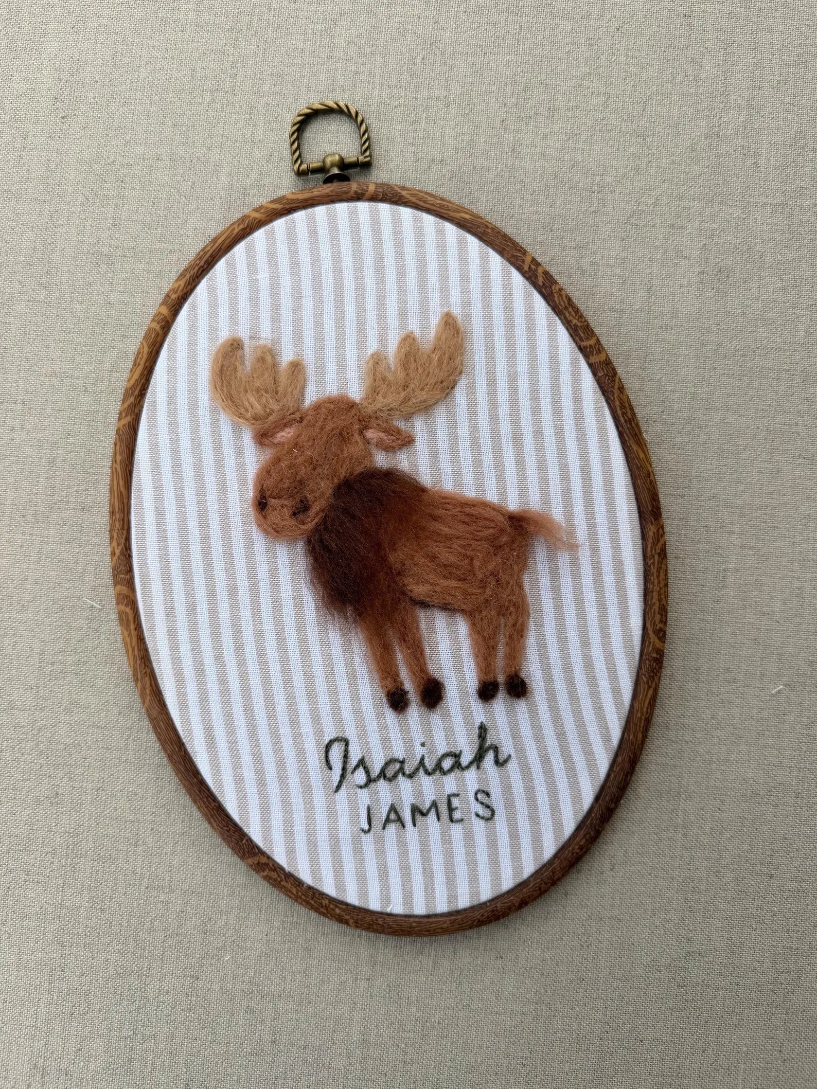 Felted moose design with 'Isaiah James' text in a wooden hoop on a striped fabric background.