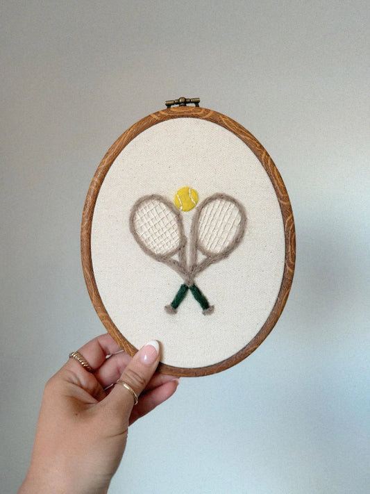 Embroidery hoop with tennis rackets and a ball design, held by a hand against a neutral background.