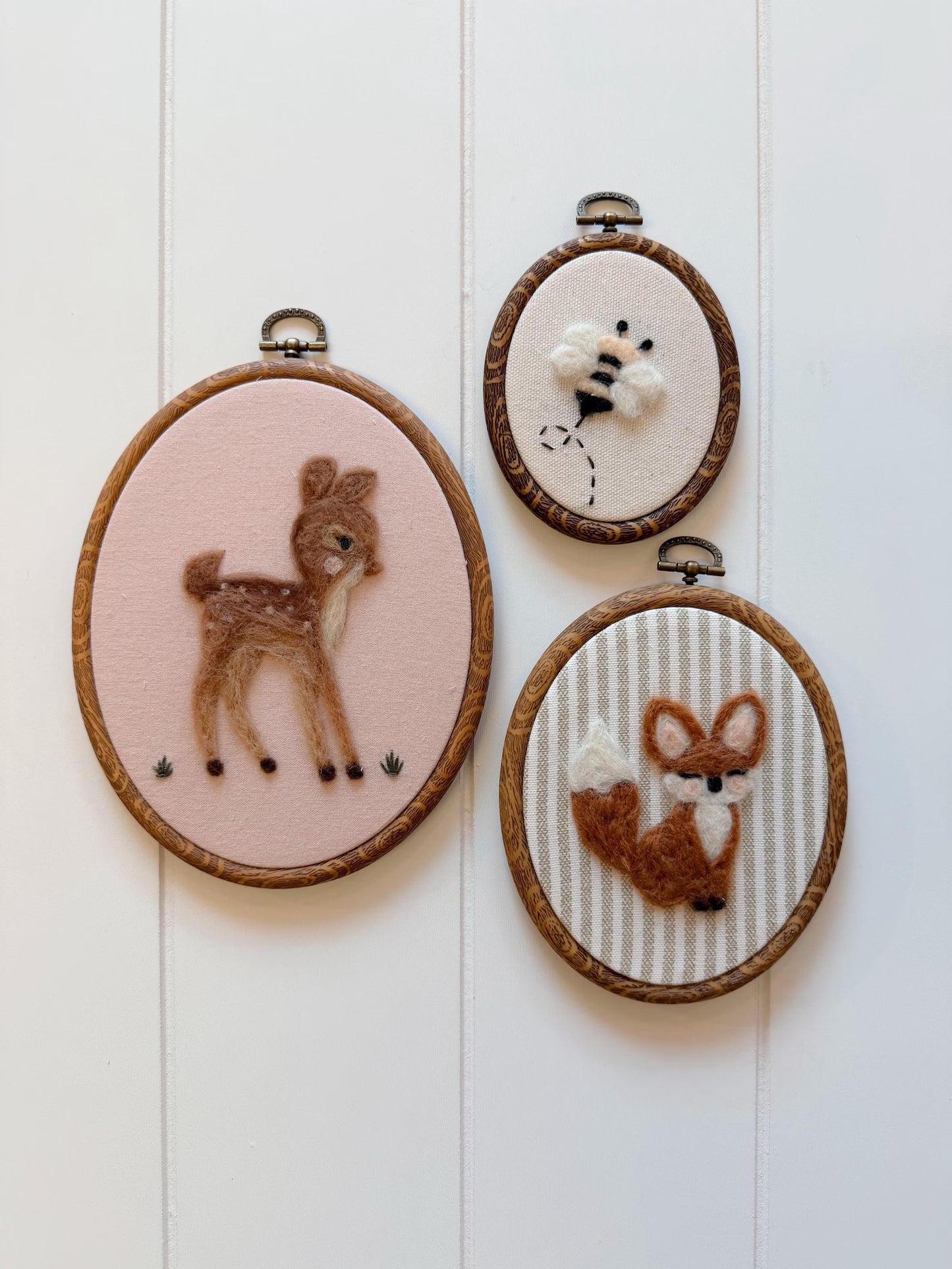 Three embroidered hoop art pieces featuring a deer, bee, and fox on a white wooden background.