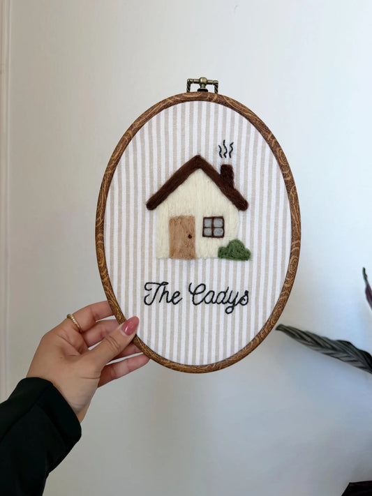Embroidery hoop with a house design and 'The Cady's' text held by a hand against a white wall.