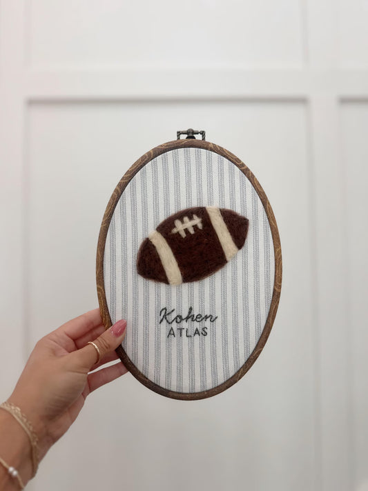 Embroidery hoop with a football design and text held by a hand against a white background